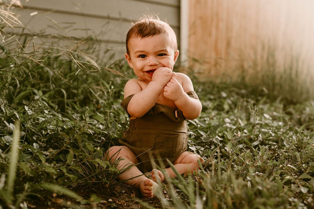 Baby in brown romper sitting in garden greenery with joyful expression.