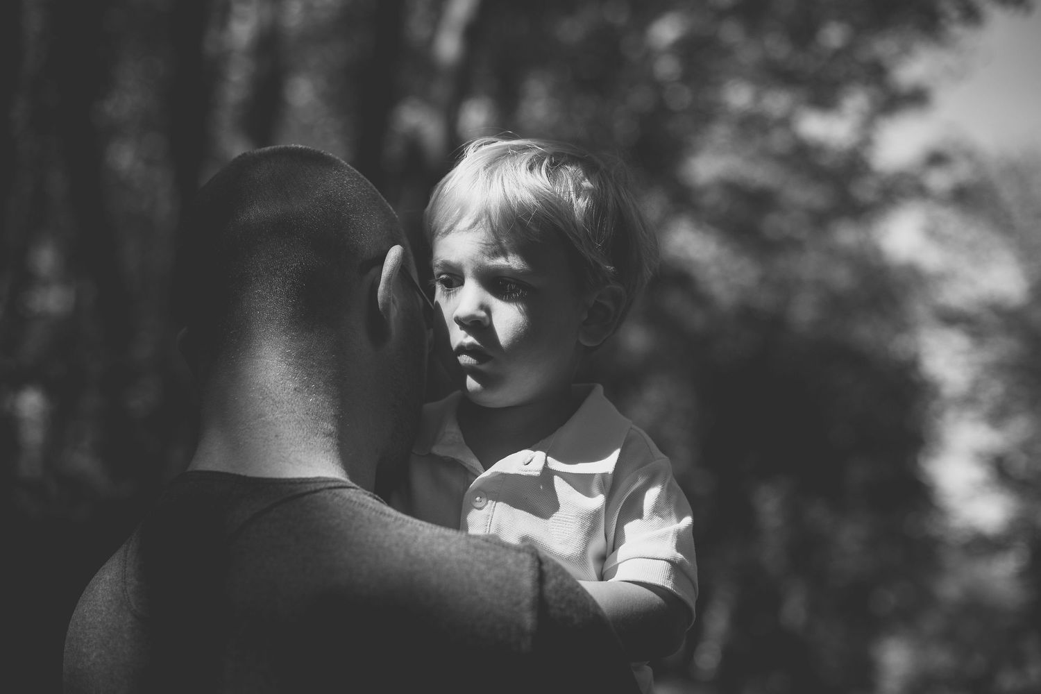 A black and white photo shows a child being held up outdoors with trees in the background.