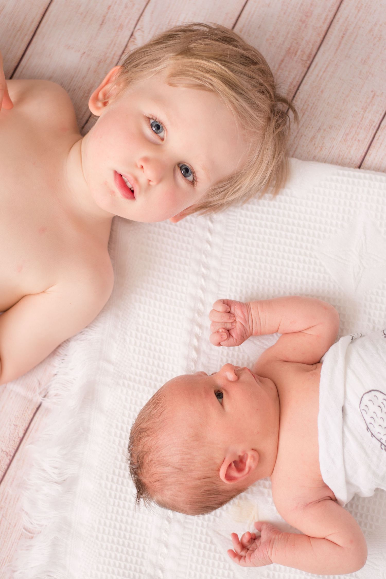 newborn baby looking over at shirtless big brother who is laying near him and looking into the camera.