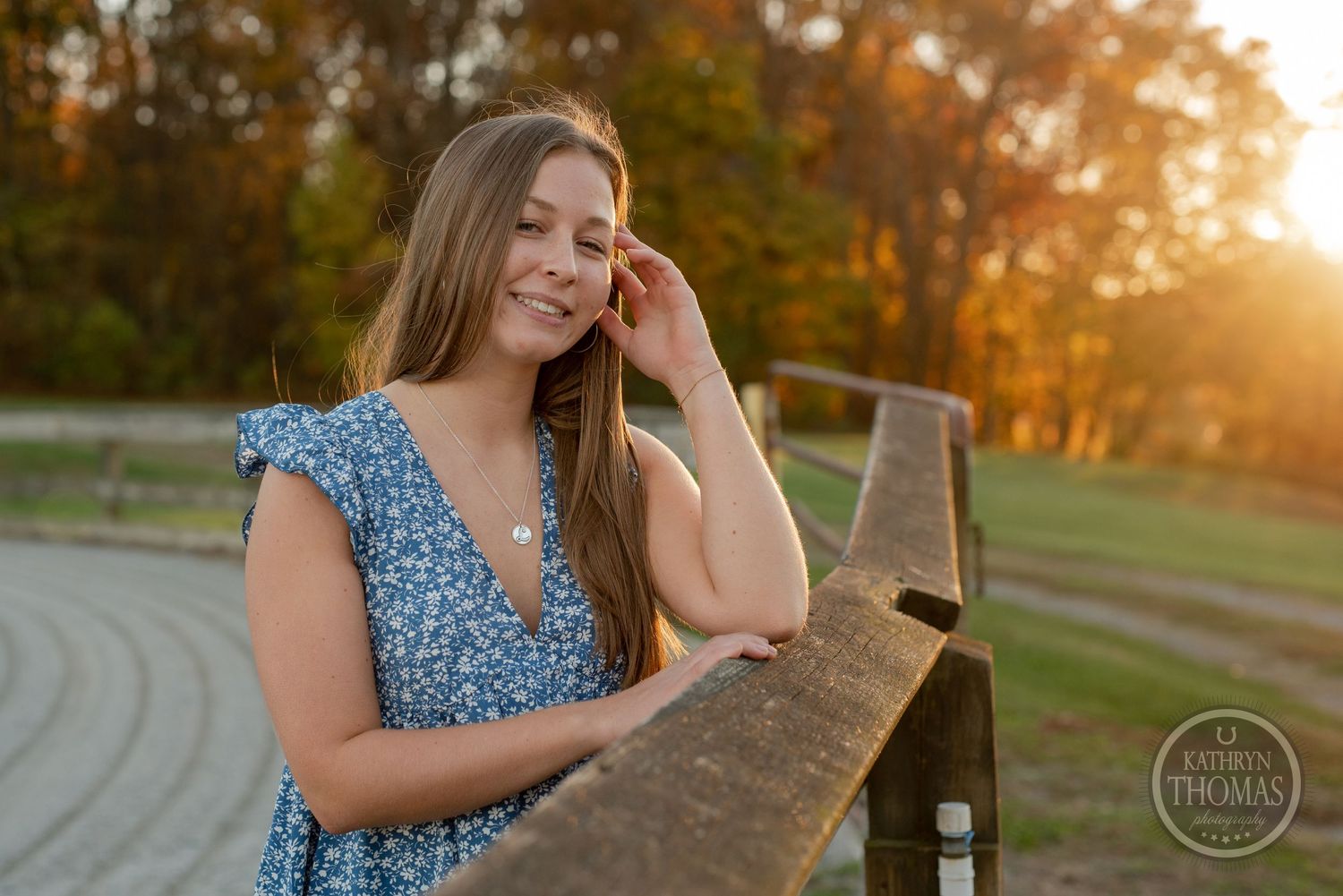 Person in blue floral dress poses by wooden fence during golden sunset in countryside setting.