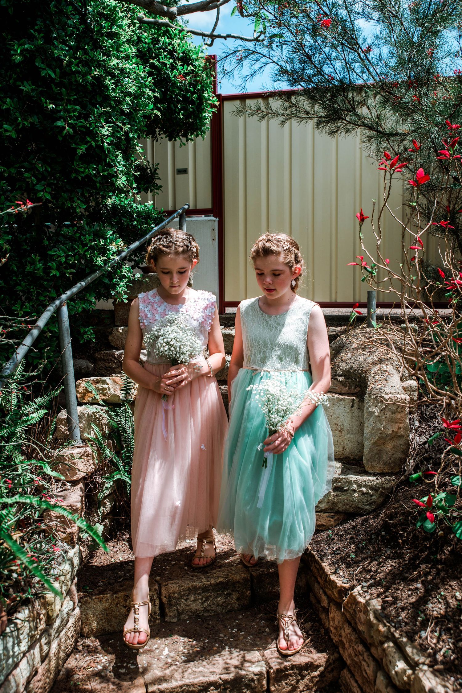 Two people in pastel formal dresses standing on garden steps with flowering vines.