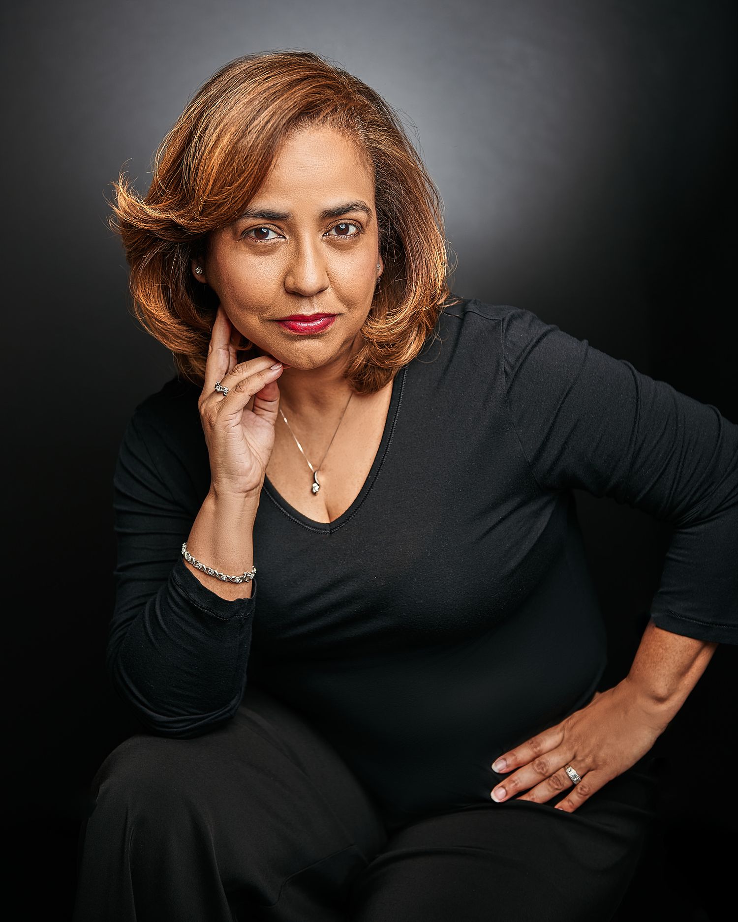 Elegant studio portrait of a woman in black long-sleeve top seated against dark background.