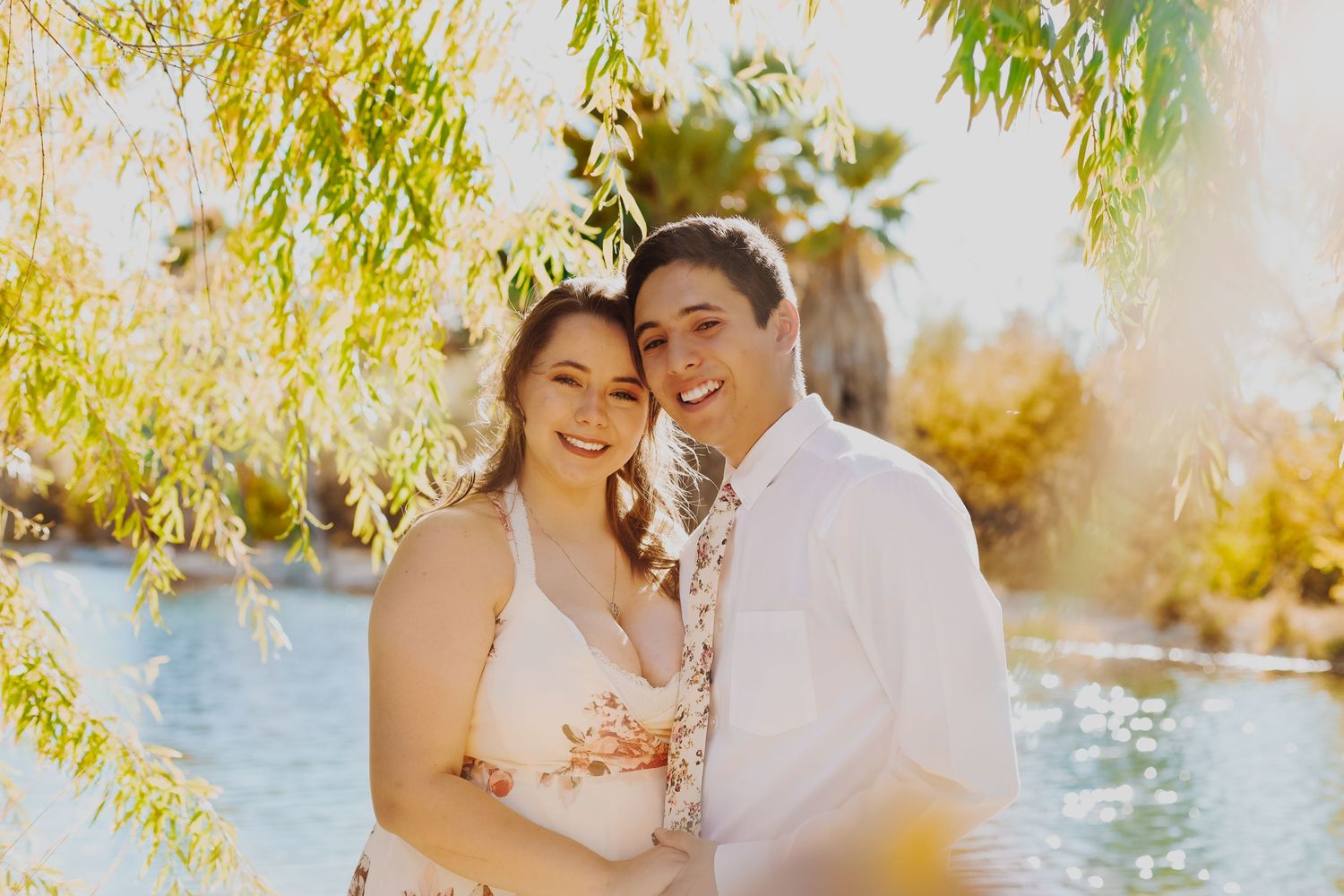 Sweet couple embraces under willow branches near a sunlit pond during romantic outdoor photoshoot.