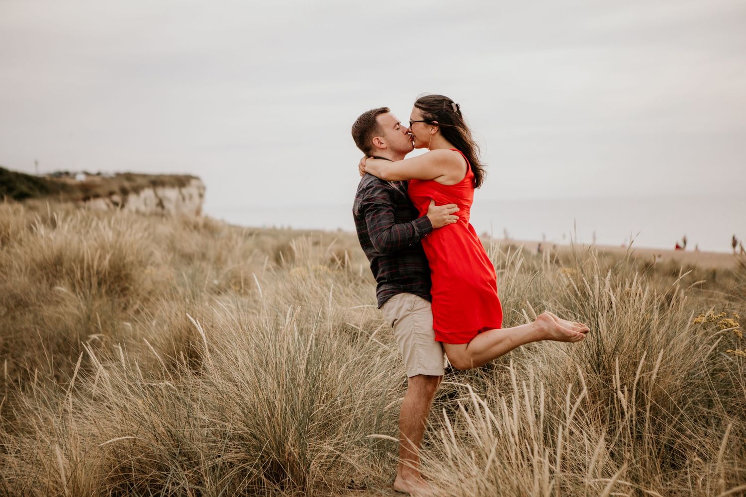 A romantic couple embraces in tall beach grass with cliffs in the background during golden hour.
