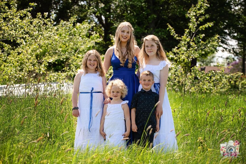 A group in white dresses and formal attire poses together in a sunny meadow.
