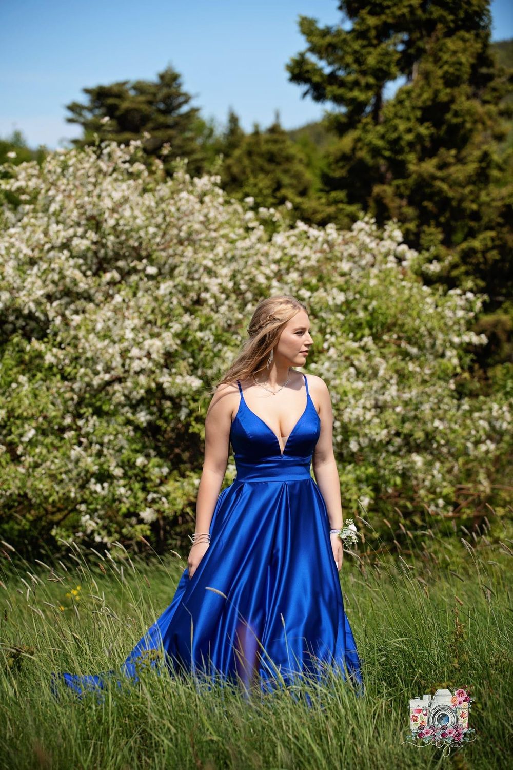 A woman in a flowing royal blue gown poses elegantly against flowering bushes and evergreen trees.