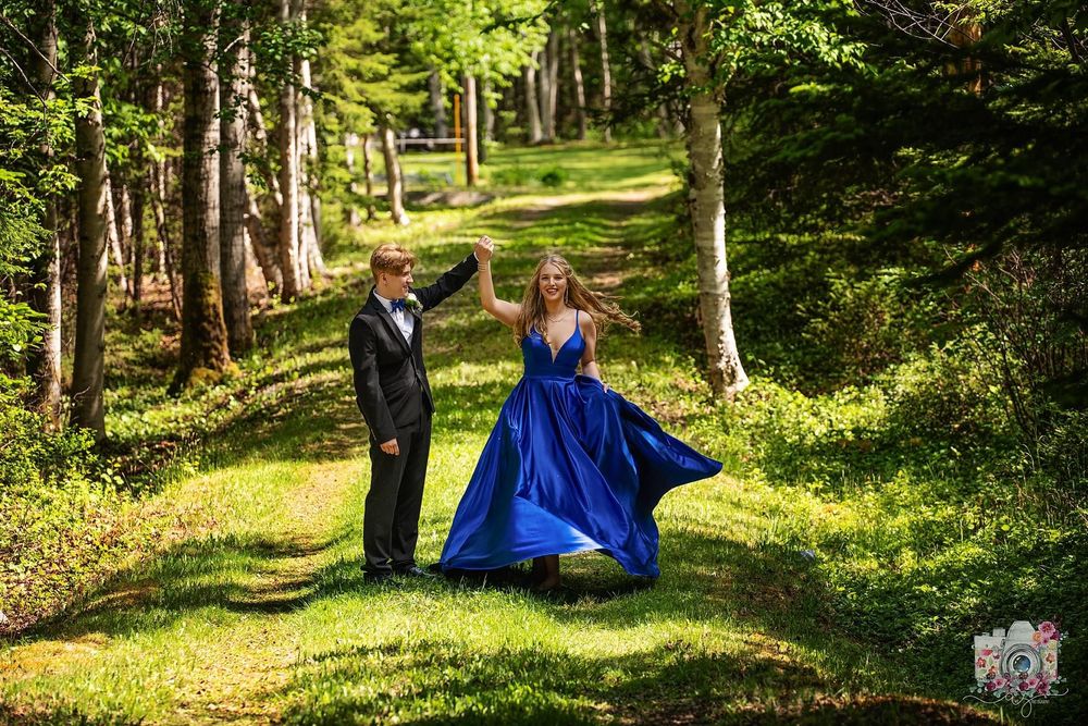 A couple in formal wear poses on a sunlit forest path, with a blue gown flowing in the breeze.