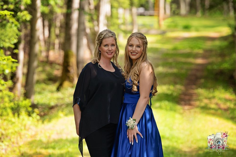 Two people stand together on a wooded path, one wearing a royal blue gown.