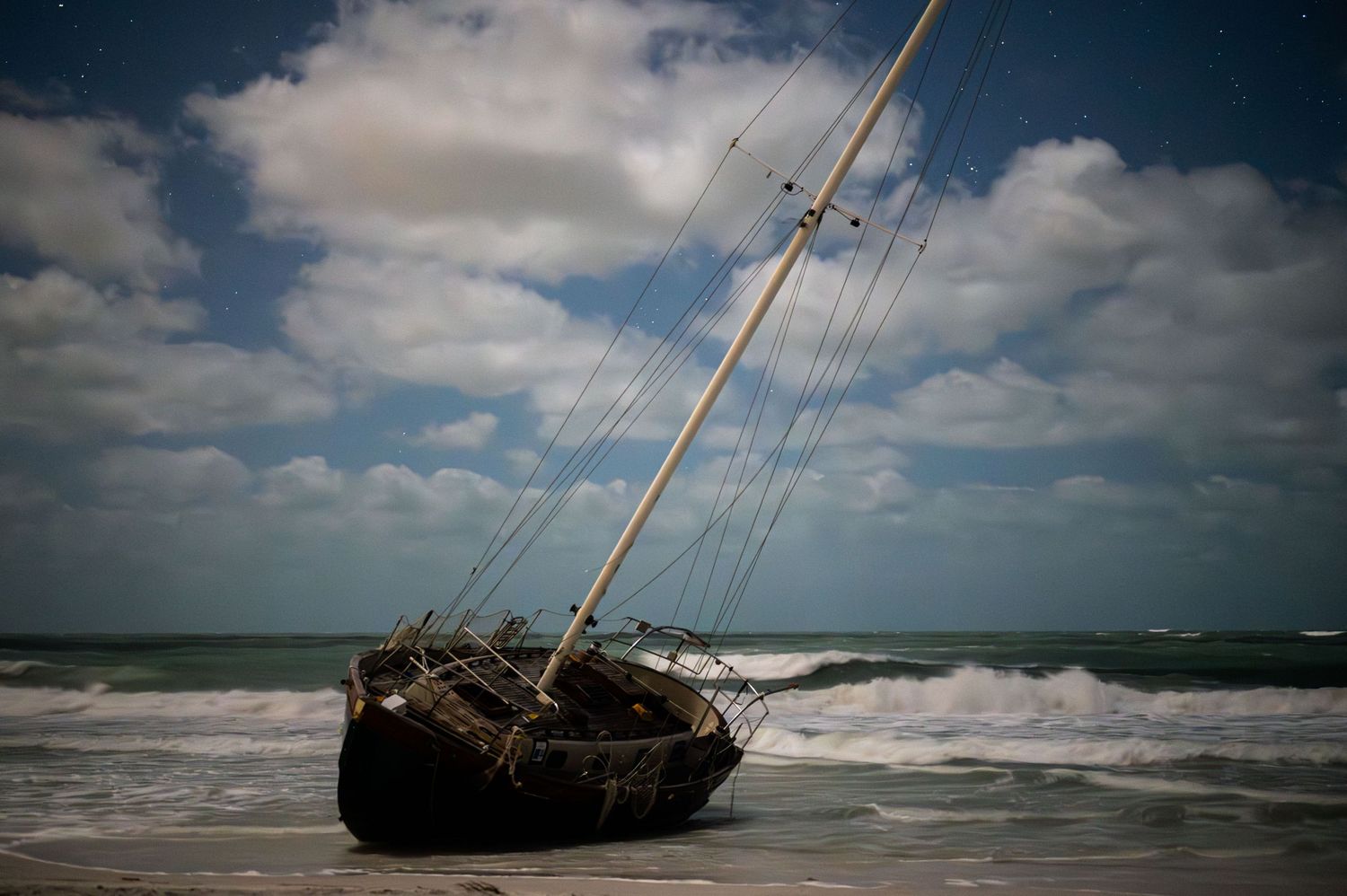 A shipwrecked sailboat lies stranded on a stormy beach with dramatic cloudy skies.