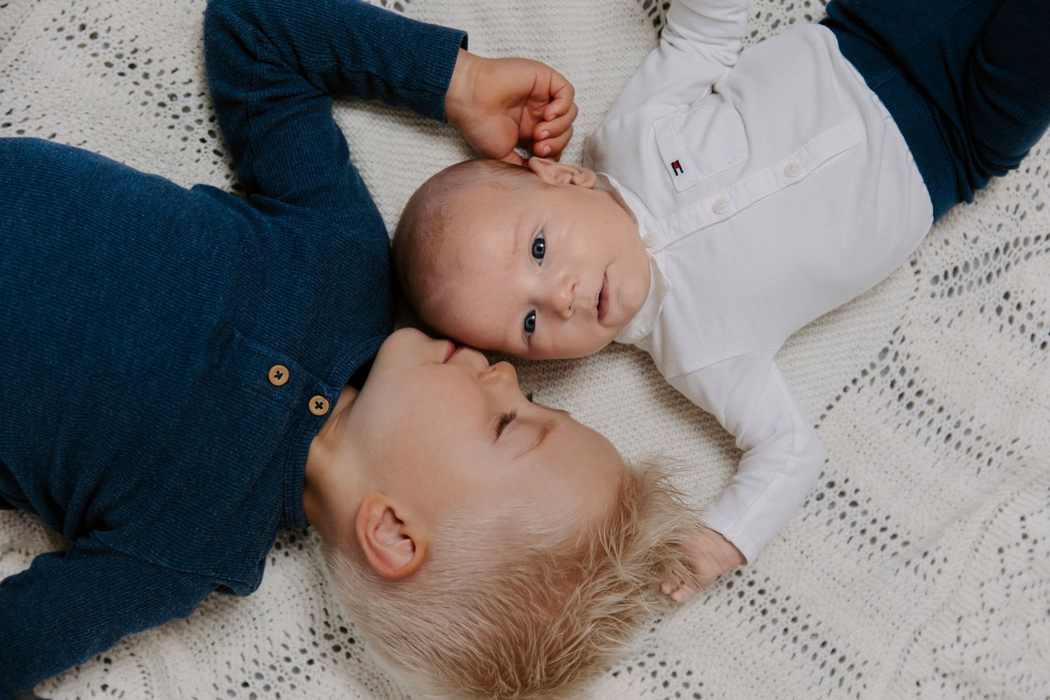 To babyer ligger med hovederne mod hinanden på et hvidt tæppe, hvor den ene rækker ud mod sit ansigt.