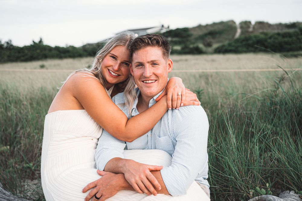 Couple embraces while sitting in coastal grass during golden hour photoshoot.