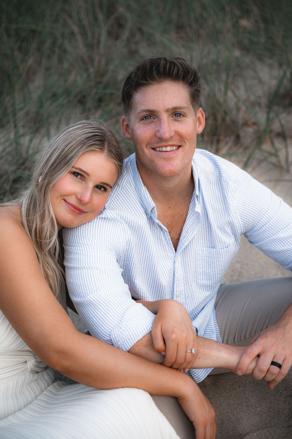 Couple relaxes together on beach while posing for casual engagement photos.