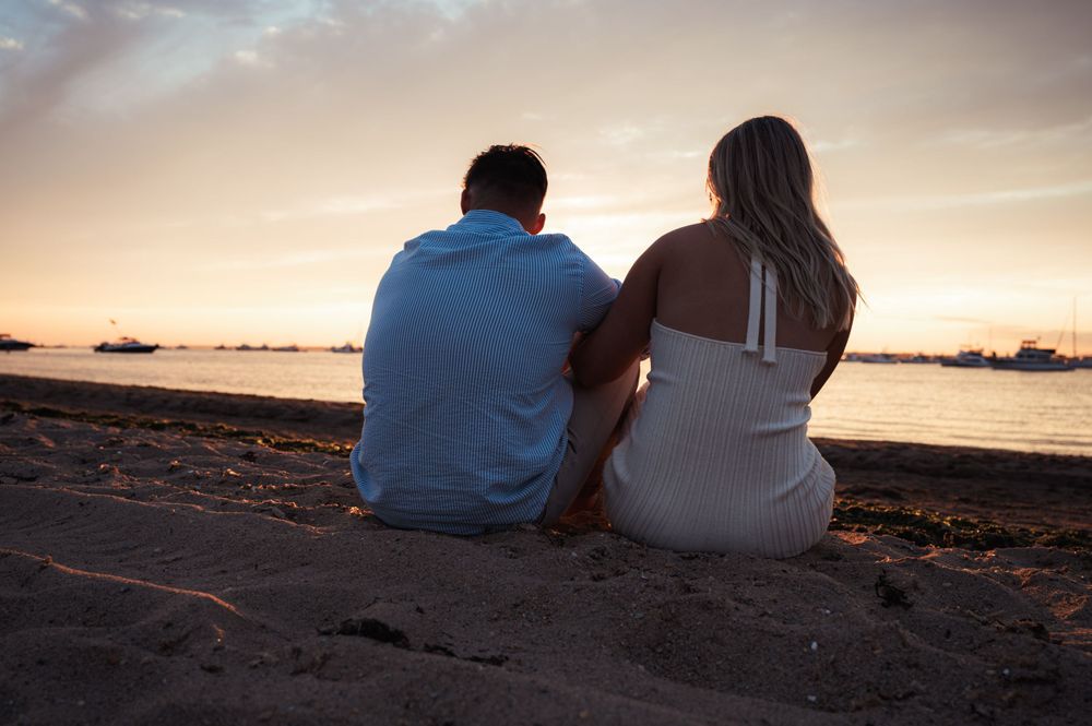 Silhouetted couple sits together on beach at sunset watching the horizon.