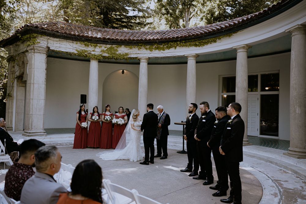 Wedding ceremony taking place in an elegant columned rotunda with bridesmaids in burgundy dresses.