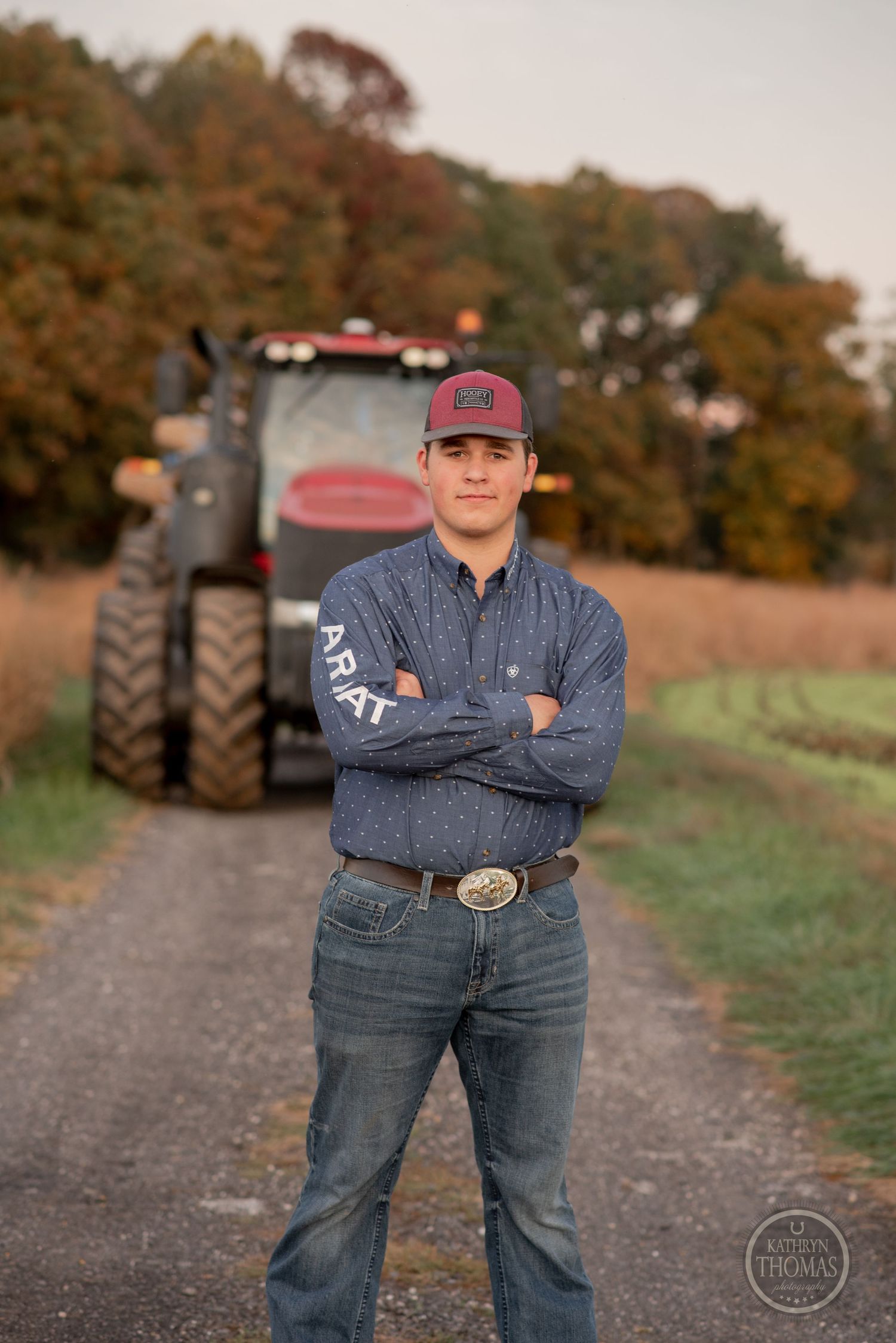 Agricultural worker stands confidently in front of large tractor with autumn foliage in background.
