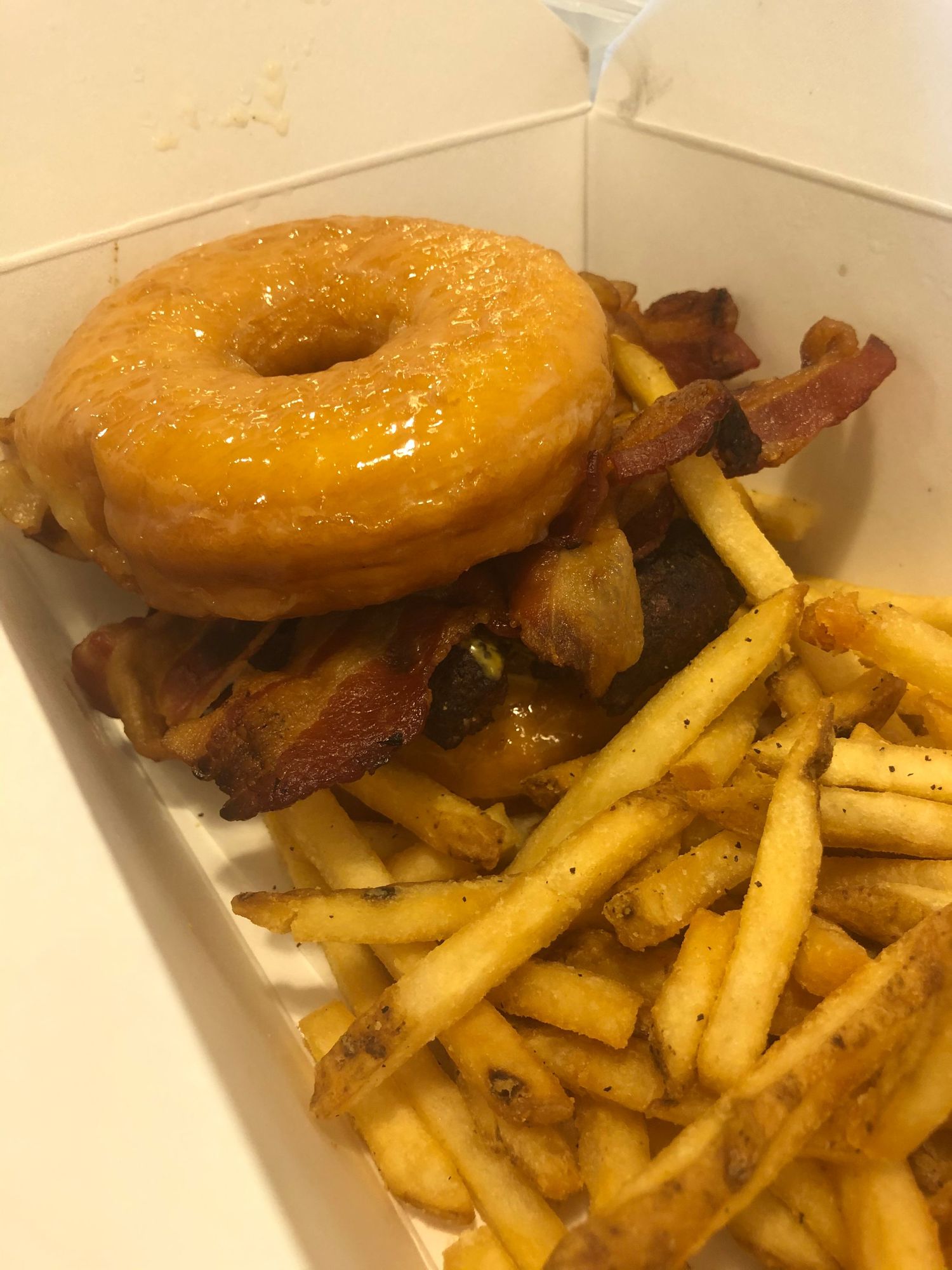 Glazed donut burger served with crispy french fries on a white takeout container.