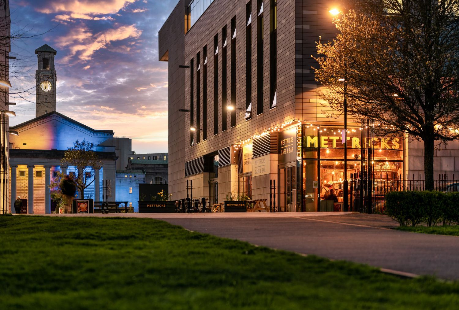 Metricks venue exterior at twilight with illuminated signage, clock tower, green lawn and dramatic sunset sky, commercial dusk photography