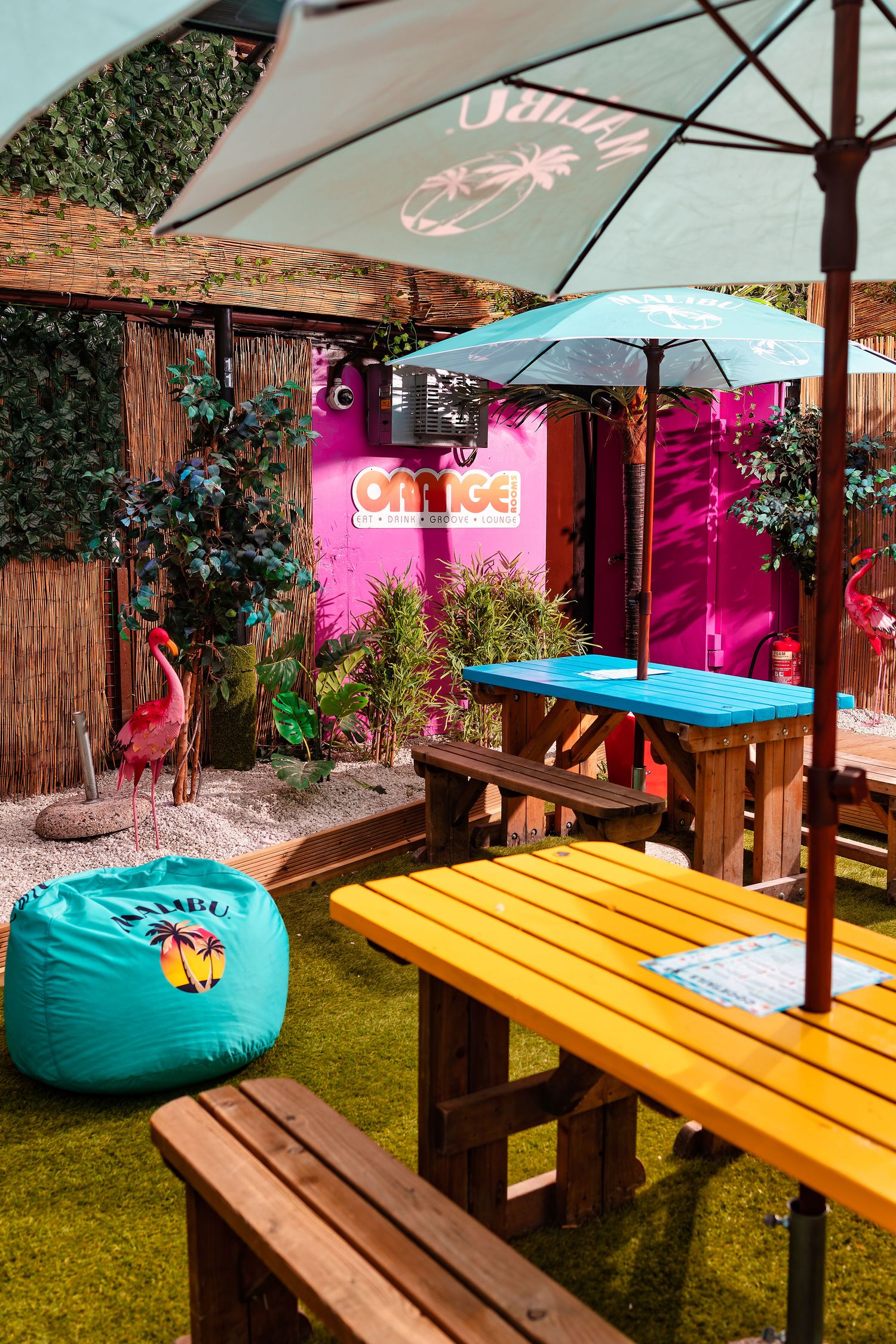 Colourful outdoor bar seating with parasols, wooden benches, pink container signage and teal beanbag on artificial grass, exterior photograp