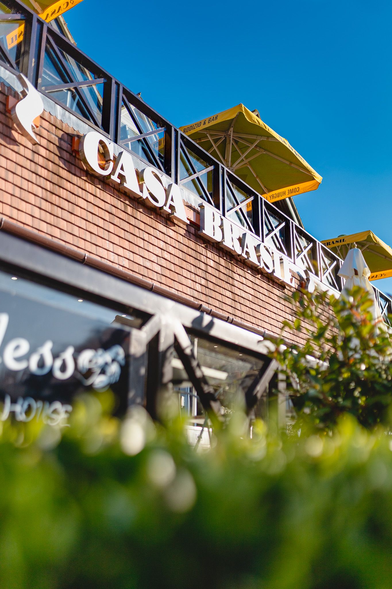 Casa Brasil building exterior with yellow canopy, brick facade and signage against blue sky with foreground greenery, architectural photogra