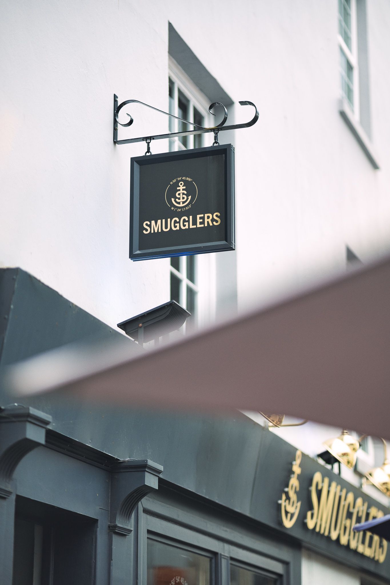 Smugglers pub hanging sign with anchor logo on wrought iron bracket against white building exterior, commercial signage photography