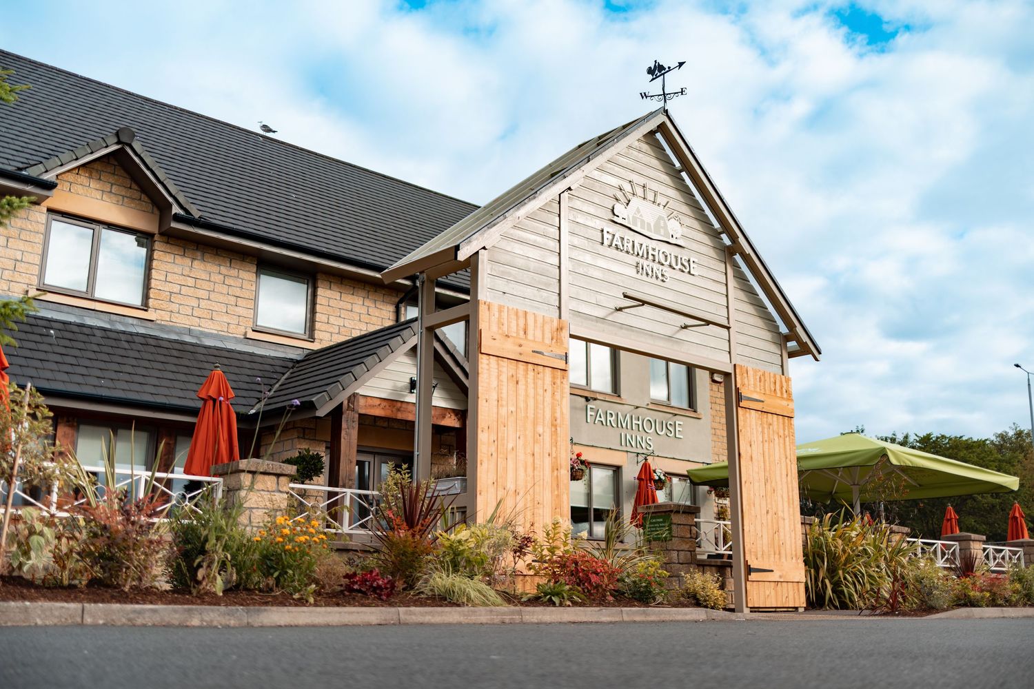 Pub exterior with stone and timber cladding, grey roof, red parasols and landscaped gardens under blue sky, commercial exterior photography