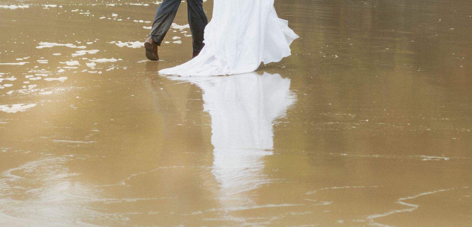 A couple in wedding attire stands on a flooded road with water reflecting their image.