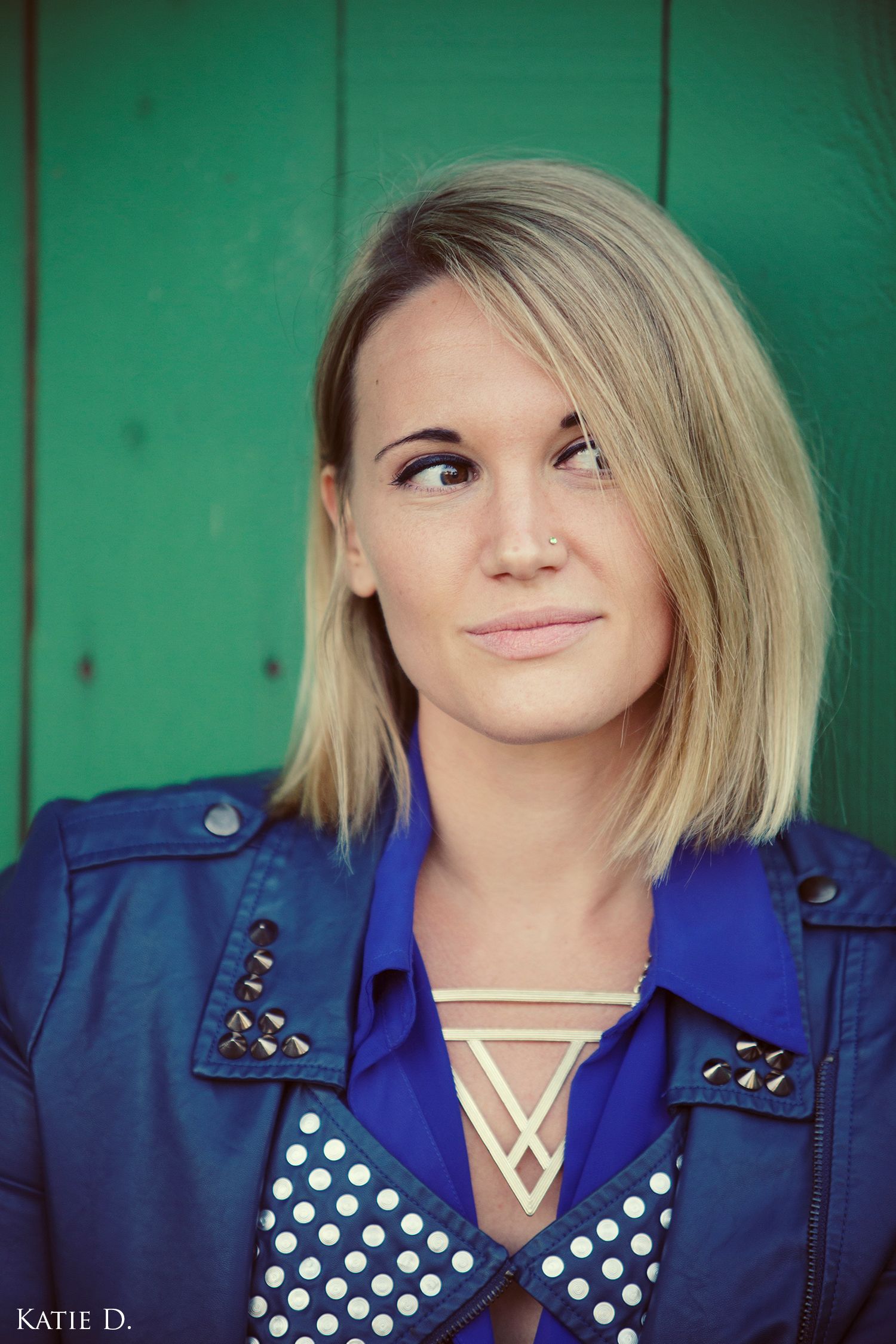 Blonde woman in blue studded leather jacket posing against green wooden wall.