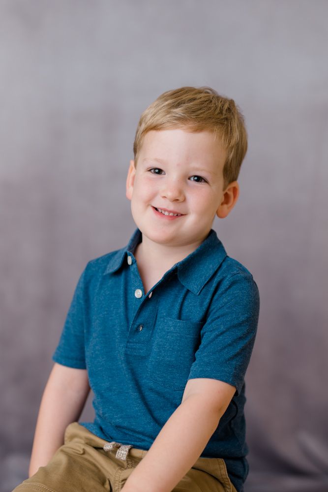 Child in a navy polo shirt and khaki pants smiles at the camera against a gray studio background.
