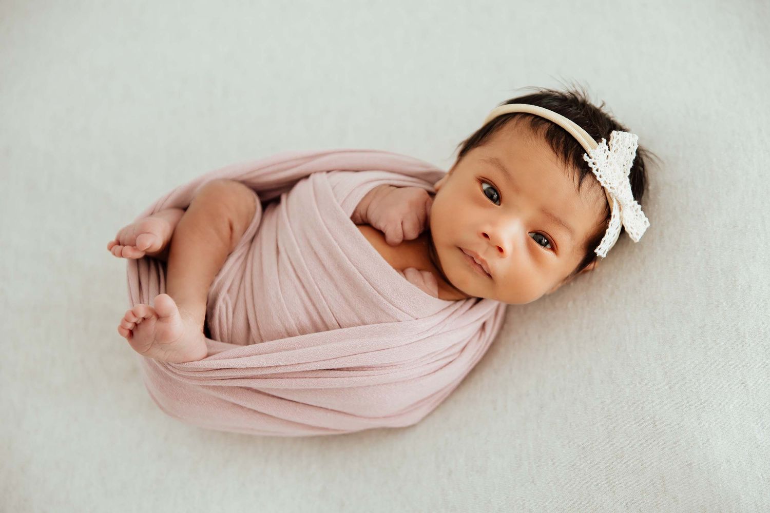 Newborn baby girl wrapped in pink swaddle with white bow headband looking up at camera.