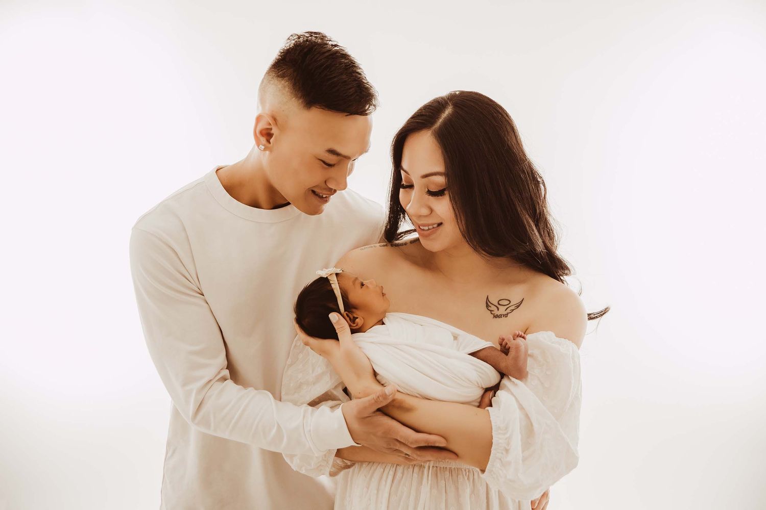 Happy couple in white outfits smiling at their newborn baby in photo studio.