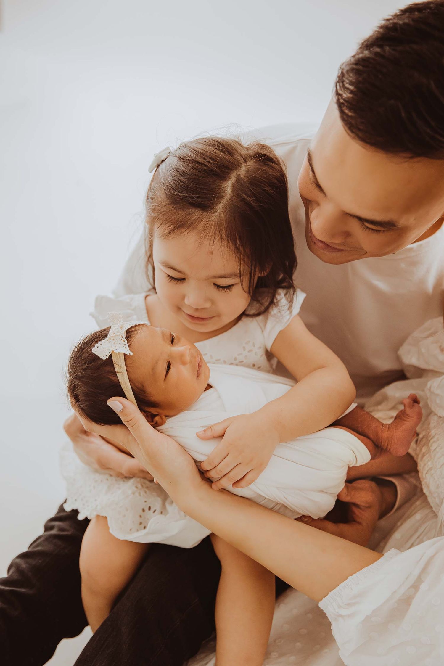 Father holds toddler daughter in white dress as she cradles newborn baby sibling in a bright studio.