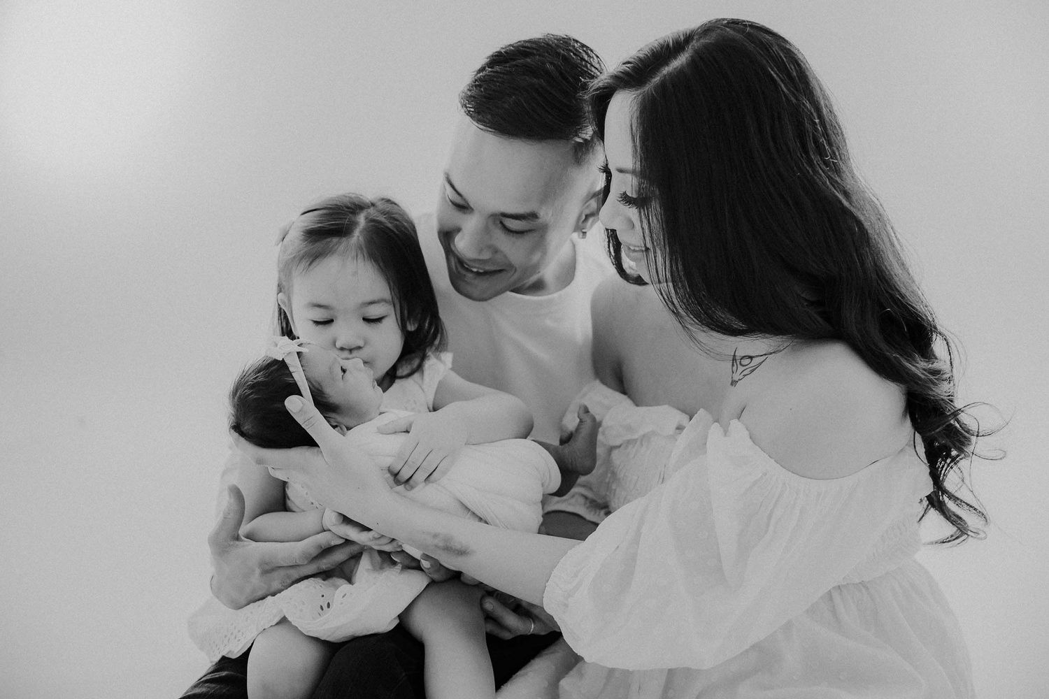 Black and white family photo of parents and toddler lovingly admiring their newborn baby in a studio.