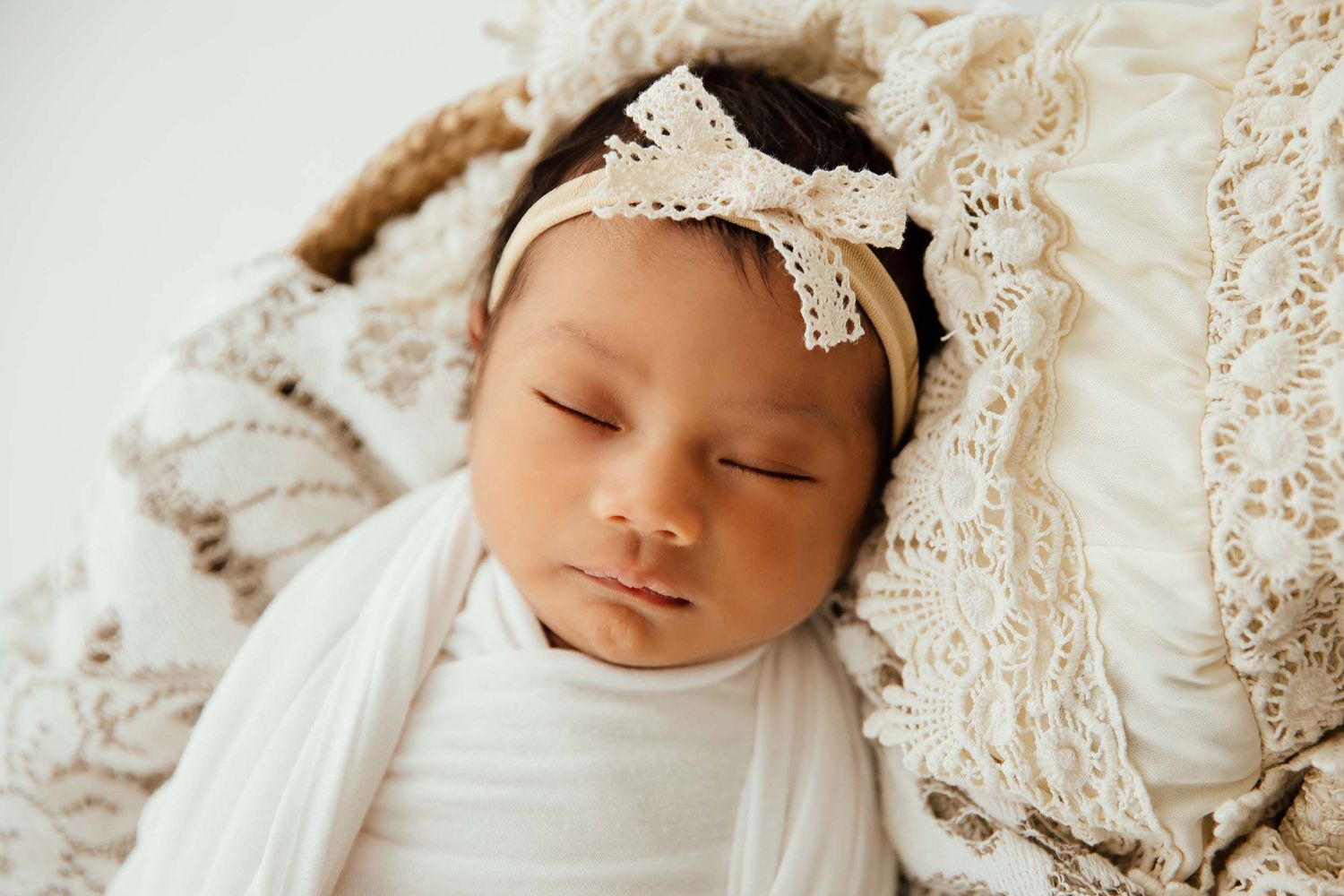 Peaceful newborn baby girl sleeping swaddled in white with a delicate lace bow headband.