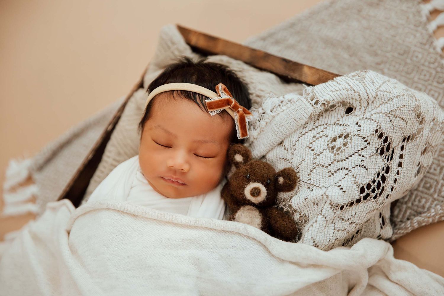 Sleeping newborn baby girl swaddled in white wrap beside a teddy bear in a wooden box.