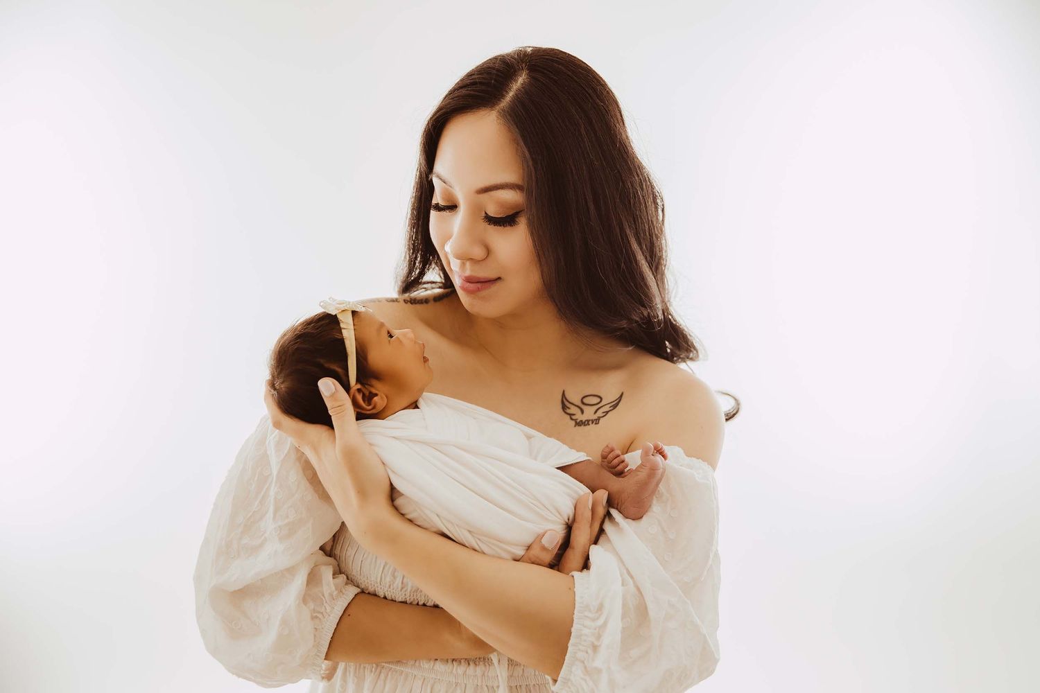 Mother in white lace dress gazes lovingly at her swaddled newborn baby in a bright studio.