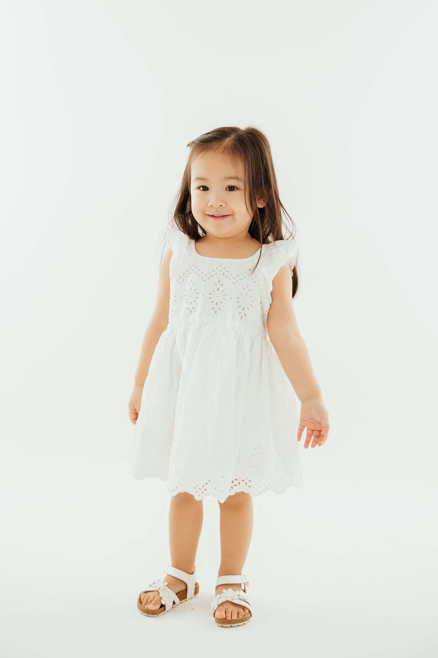 Adorable little girl in white eyelet dress and sandals smiling in studio.