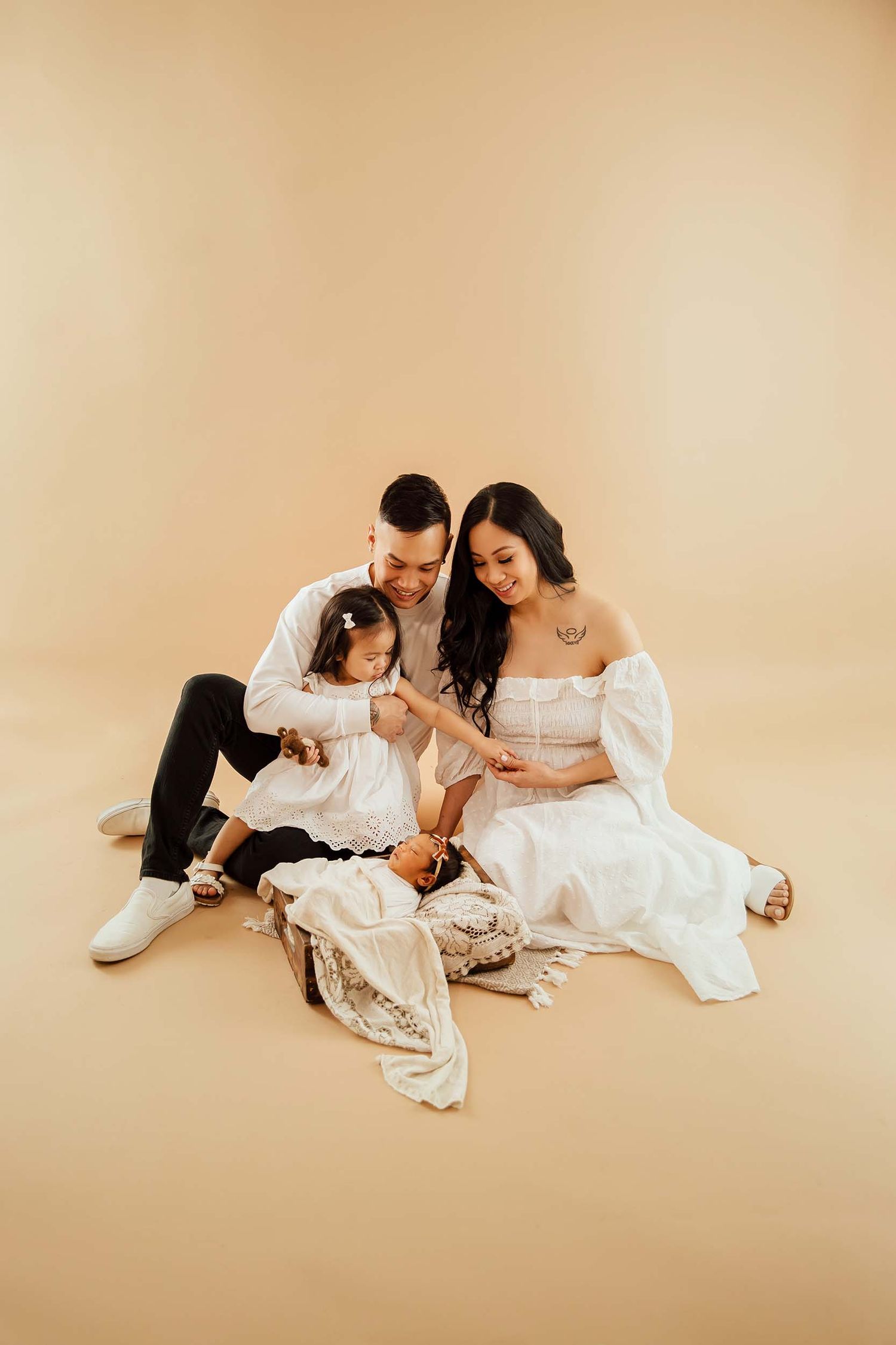 Family of three in white outfits sit together on beige backdrop admiring their newborn baby.