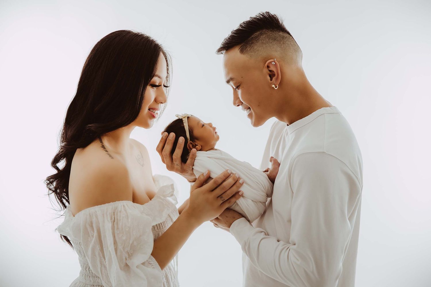 New parents in white outfits tenderly holding their newborn baby in studio.