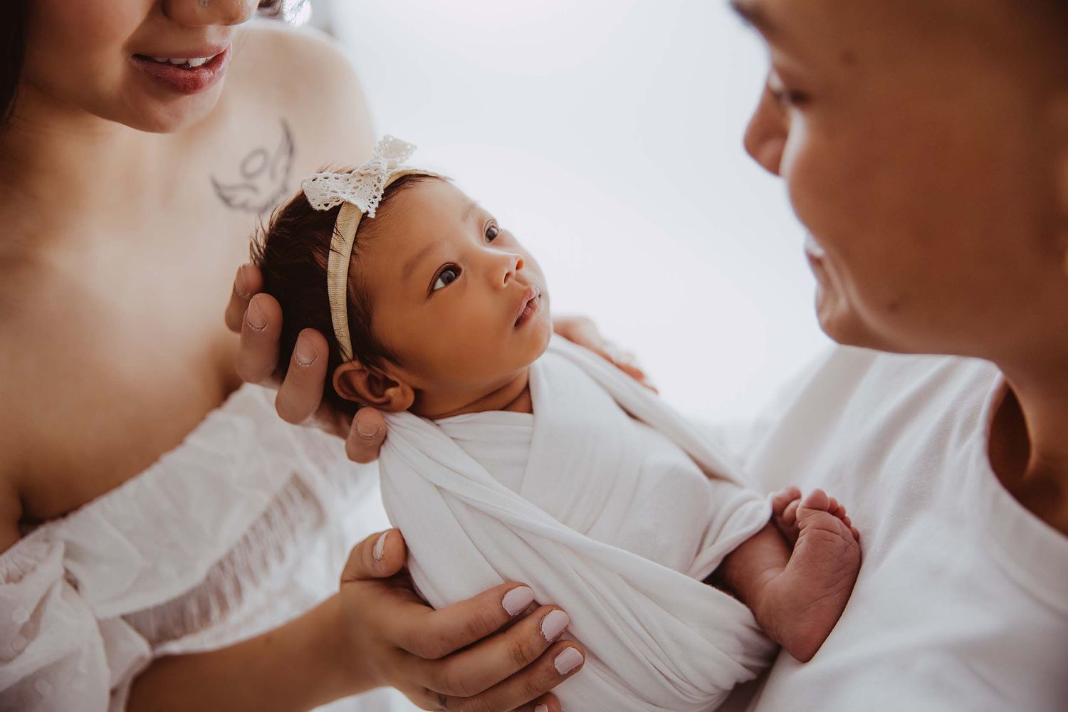 Close-up of newborn baby with headband being held by two women in white.