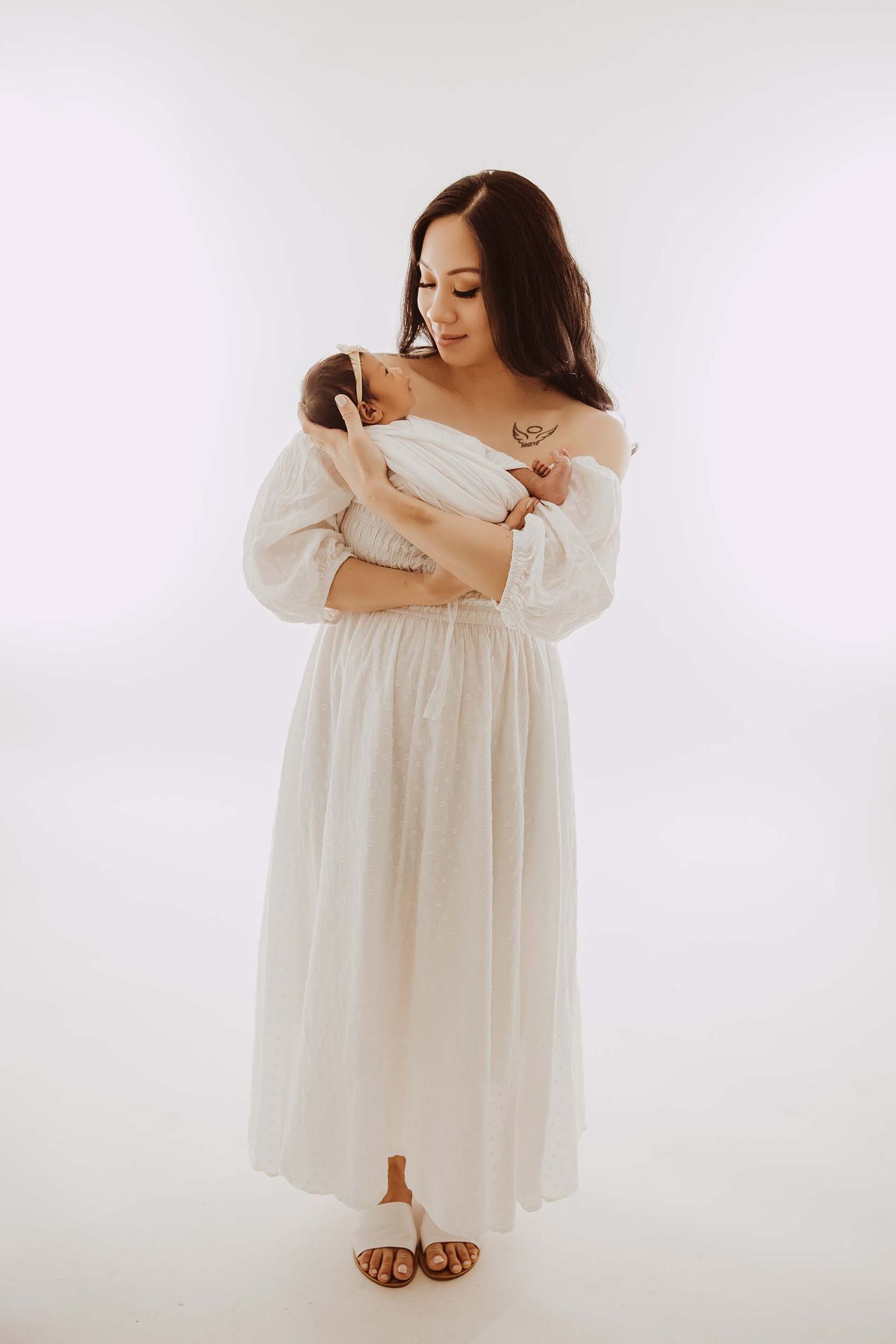 Mother in white off-shoulder dress lovingly cradling her newborn baby in studio.