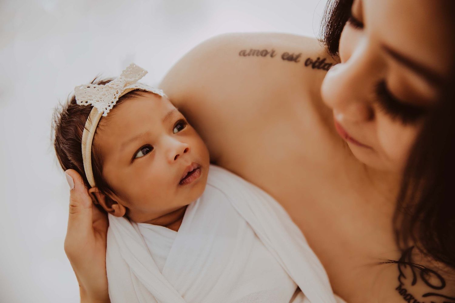 Mother with tattoo gently holding newborn baby with floral headband close to chest.
