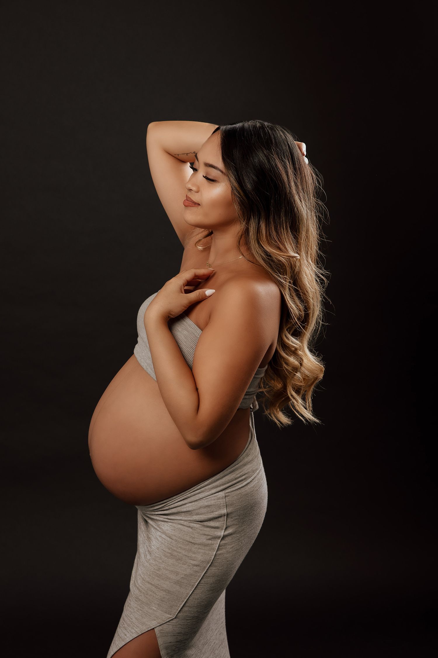 Pregnant woman in gray wrap dress poses sideways on dark background with hand raised behind her head in a Vancouver studio photoshoot.
