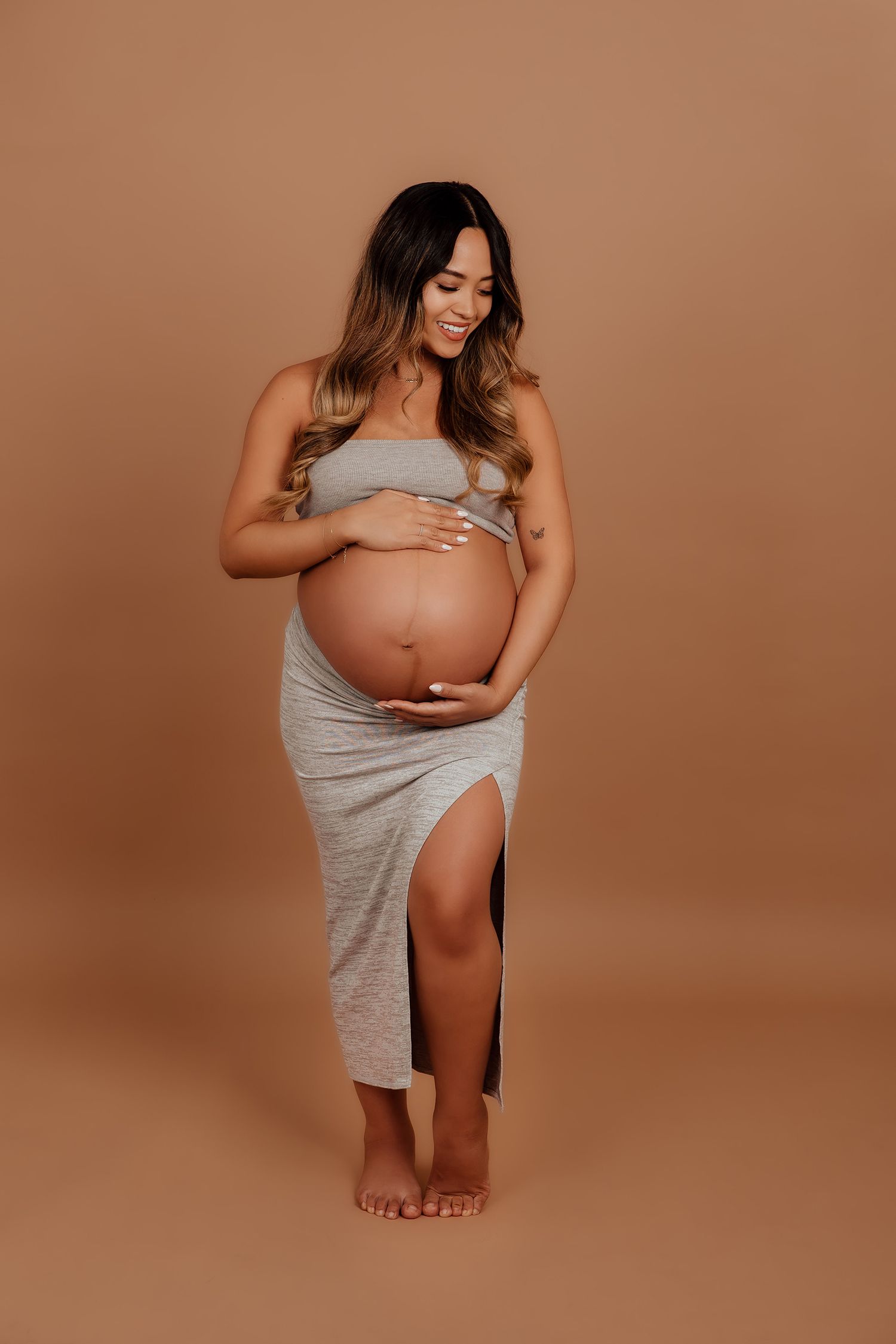 Pregnant woman in gray tube dress cradling baby bump on beige studio background in a Vancouver studio photoshoot.