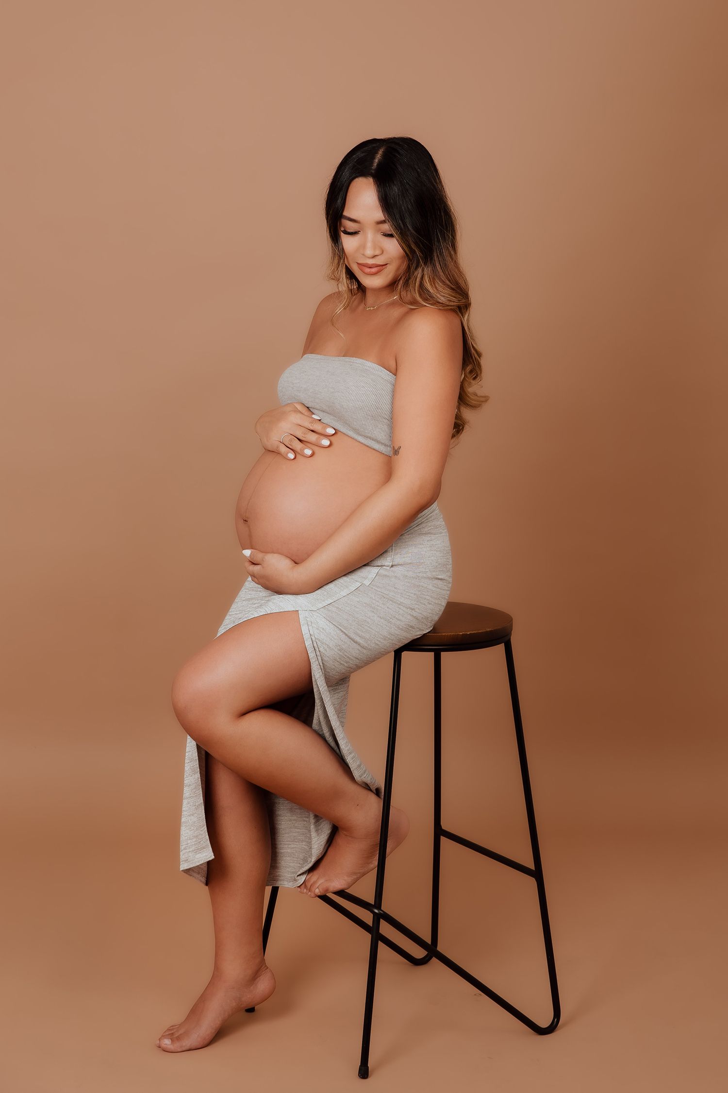 Pregnant woman in gray wrap dress seated on stool cradling bump on beige background in a Vancouver studio photoshoot.
