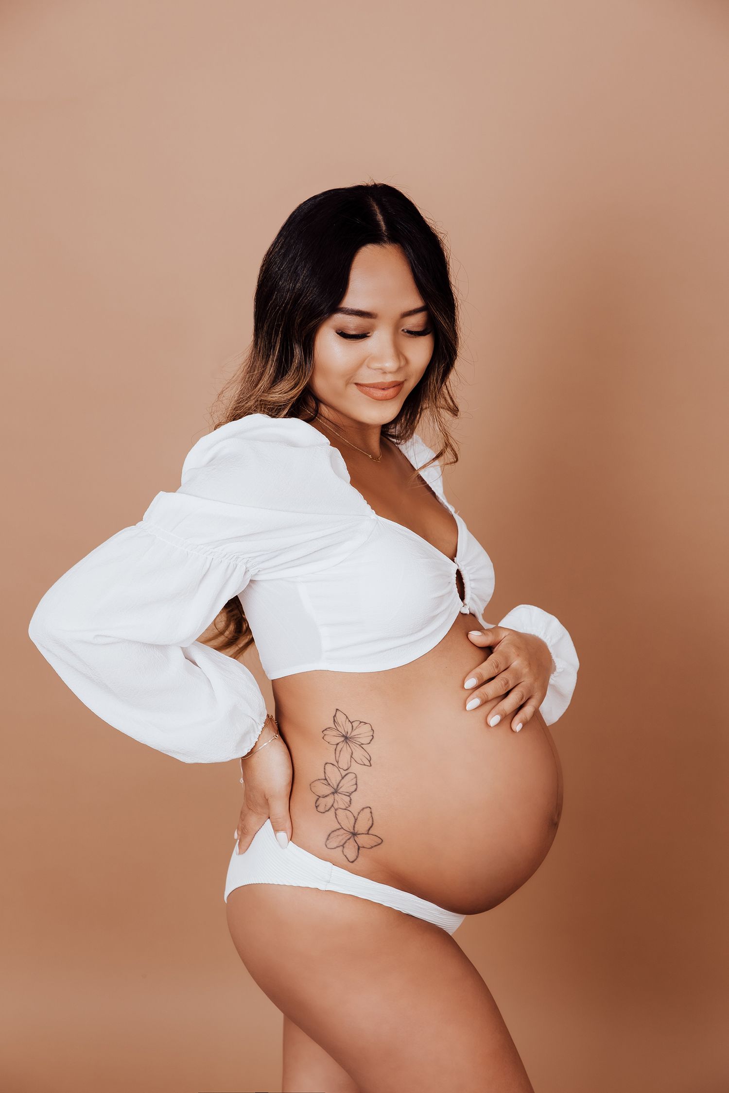 Pregnant woman in white lingerie and open blouse showing floral tattoo and baby bump in a Vancouver studio photoshoot.