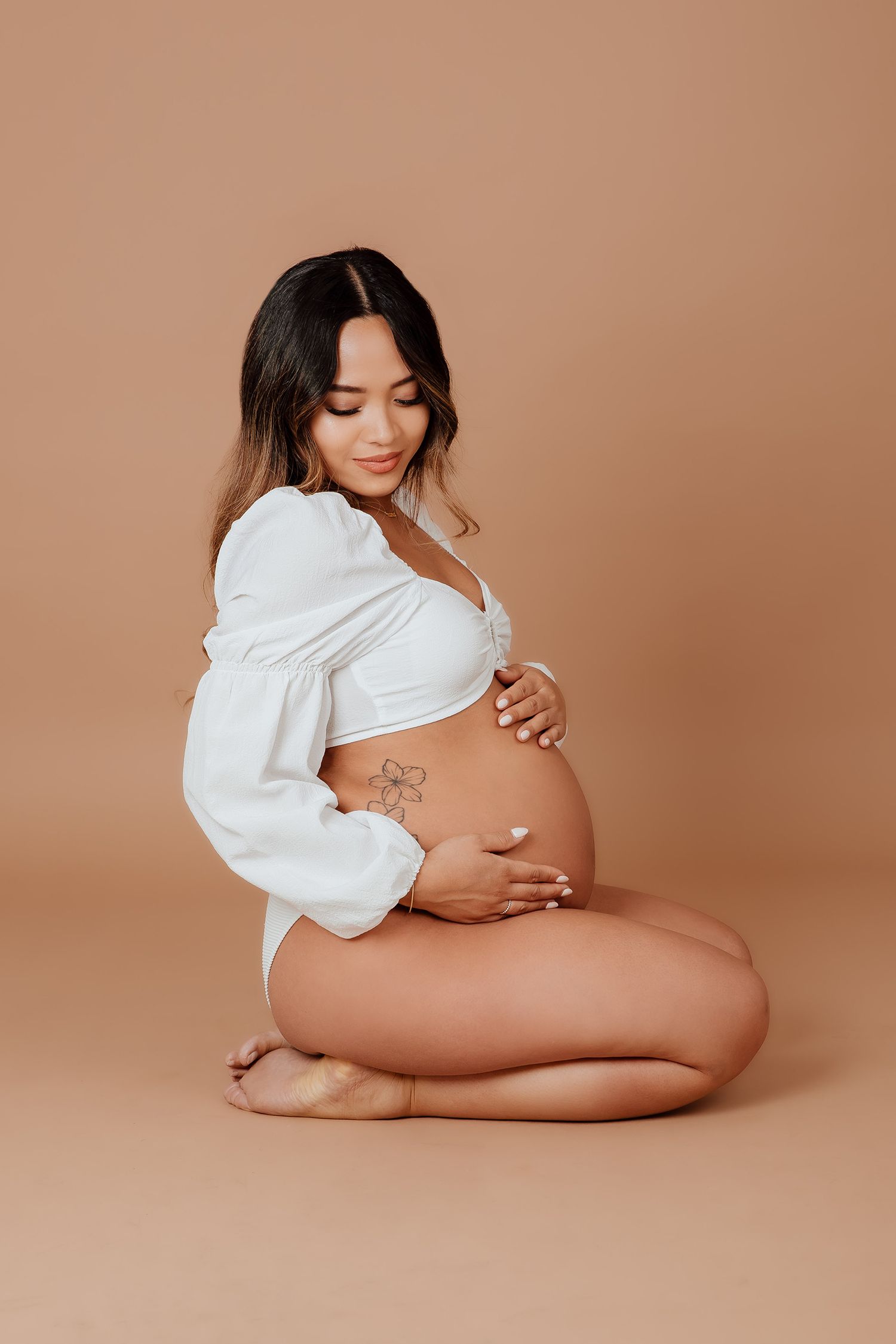Pregnant woman in white crop top kneeling on floor holding baby bump on beige background in a Vancouver studio photoshoot.
