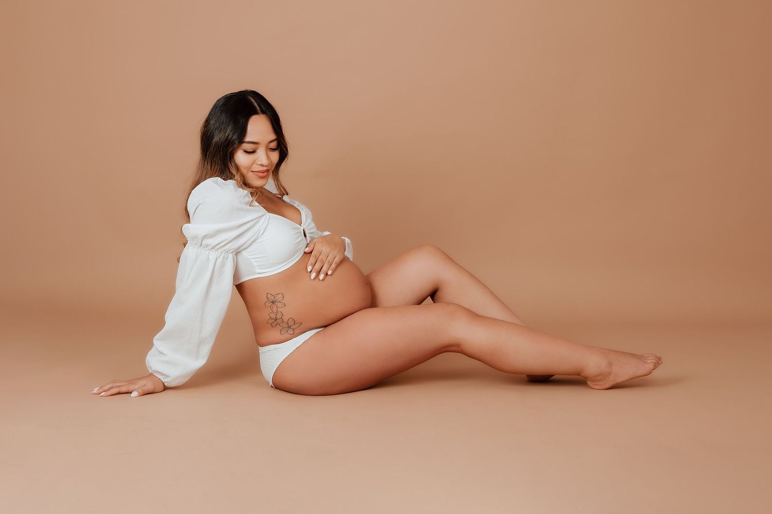 Pregnant woman in white crop top and underwear sits gracefully on beige backdrop cradling her baby bump in a Vancouver studio photoshoot.