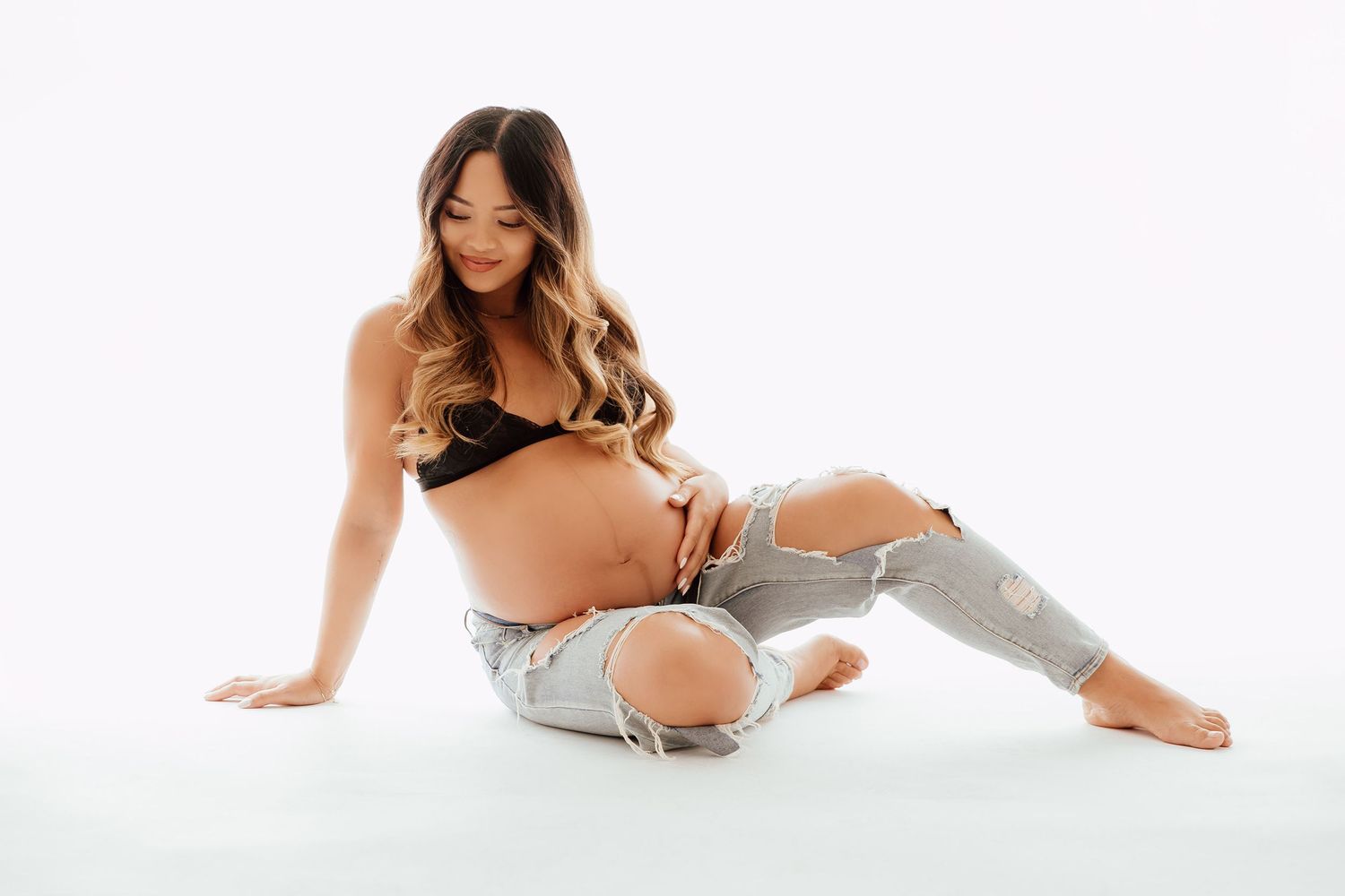 Pregnant woman in black bralette and torn jeans sits barefoot on white floor holding her baby bump in a Vancouver studio photoshoot.
