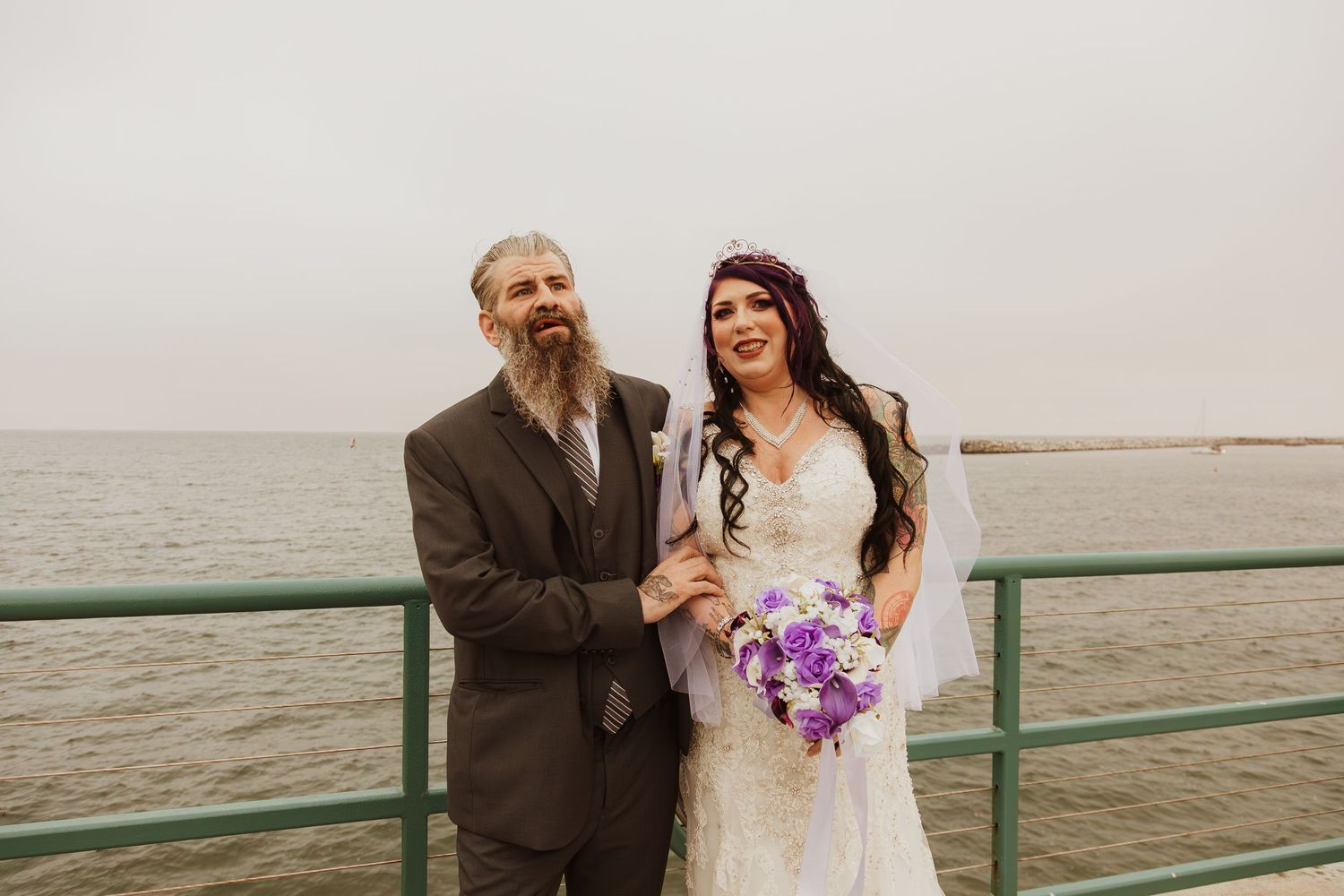 A couple on their wedding day poses by the ocean with purple flowers.