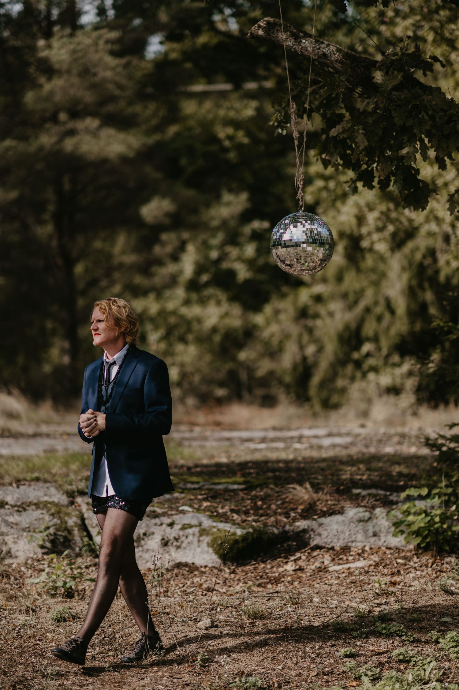 Person in blazer and shorts walks near hanging disco ball in forest setting.